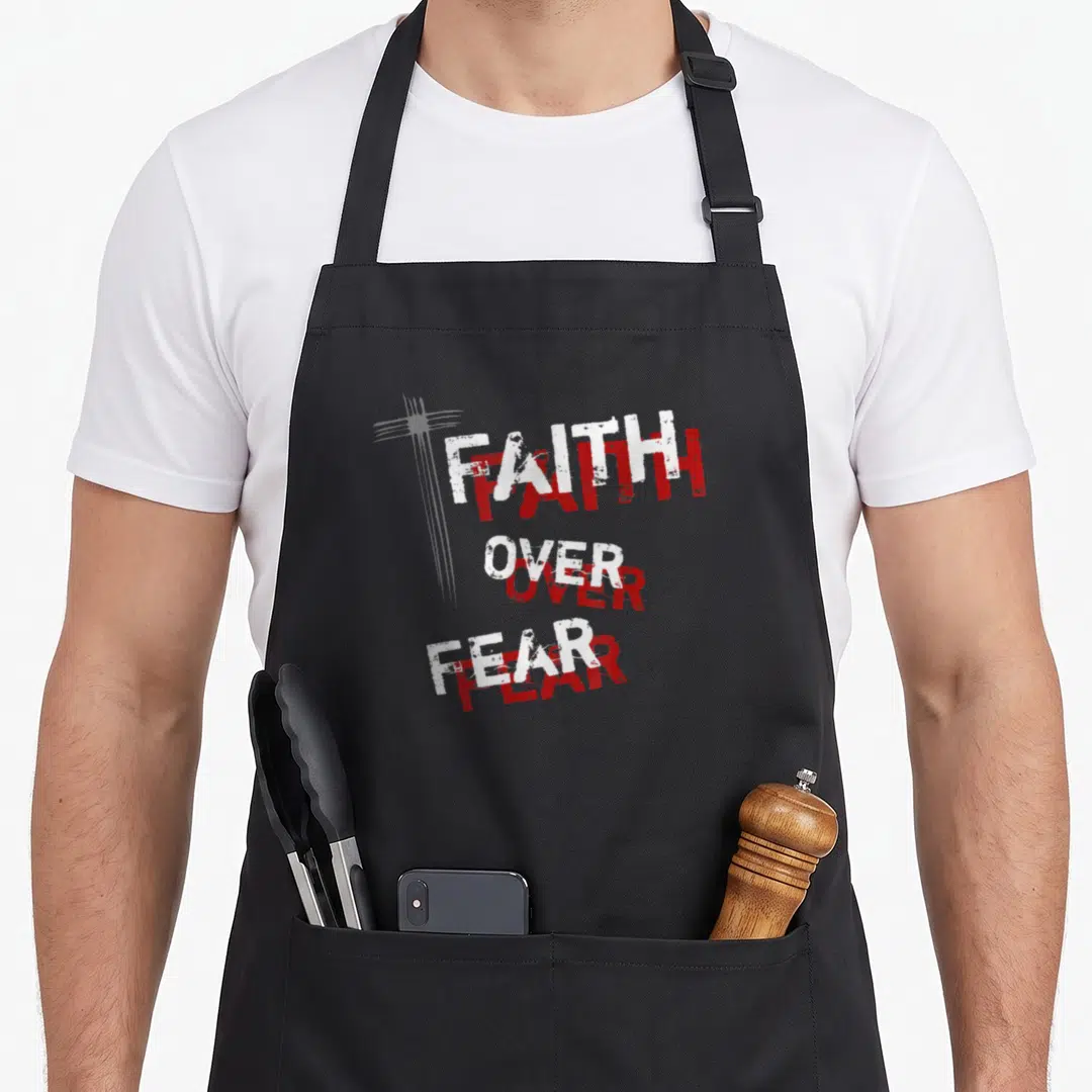 Inspirational cross over apron - Stacked FAITH OVER FEAR text in white and red distressed font with grey cross — shown on a black cotton canvas apron against a white background