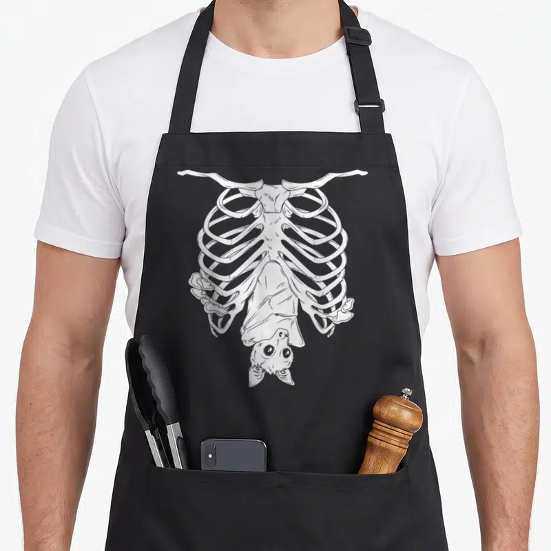 Wiccan goth apron - White line-art skeleton ribcage with a cute bat hanging upside down inside the chest cavity — shown on a black cotton canvas apron against a white background
