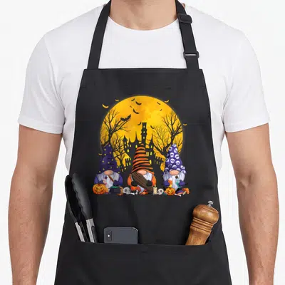 Witch fall aprons - Three cartoon gnomes in witch hats holding Halloween props before a yellow moon and castle — shown on a black cotton canvas apron against a white background