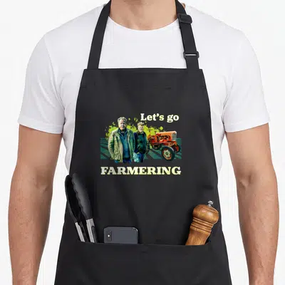 Farmering farm apron - Two men and orange tractor under Let's go text above FARMERING word in cartoon style — shown on a black cotton canvas apron against a white background