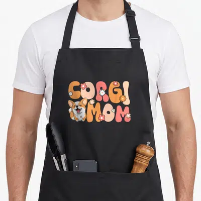 Groovy corgi aprons - CORGI MOM text in orange pink bubbles with cartoon corgi and scattered flowers — shown on a black cotton canvas apron against a white background