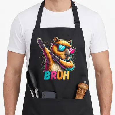 Capy meme apron - Dabbing capybara in brown with sunglasses, BRUH text in blue pink orange yellow, radiating light — shown on a black cotton canvas apron against a white background