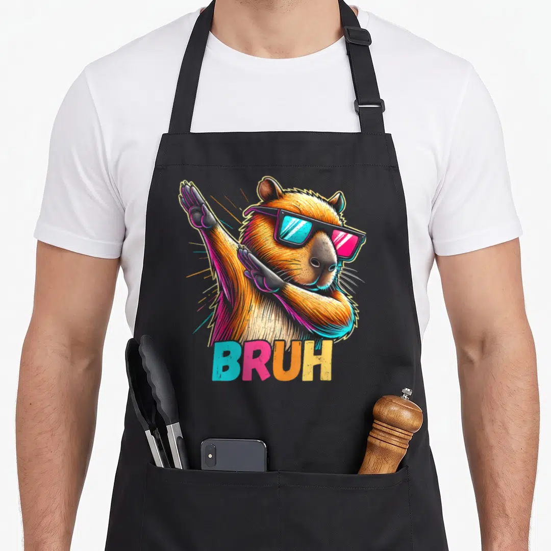 Capy meme apron - Dabbing capybara in brown with sunglasses, BRUH text in blue pink orange yellow, radiating light — shown on a black cotton canvas apron against a white background