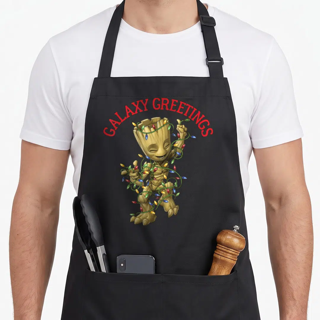 Greeting christmas apron - Groot wrapped in red green yellow blue lights under arched red GALAXY GREETINGS text — shown on a black cotton canvas apron against a white background