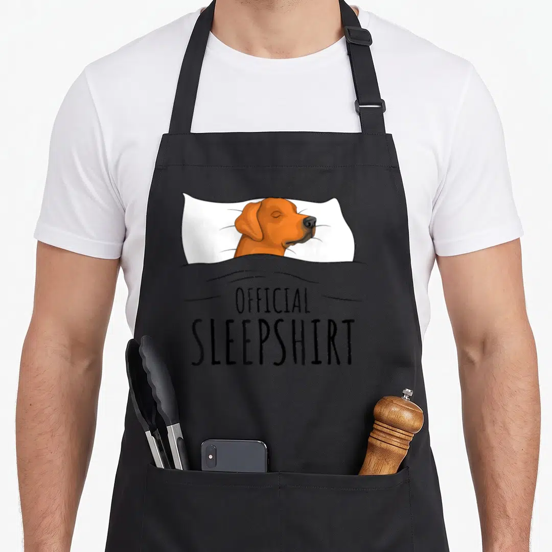 Ridgeback sleepshirt dog apron - Orange dog sleeping under grey blanket with OFFICIAL SLEEPSHIRT text — shown on a black cotton canvas apron against a white background