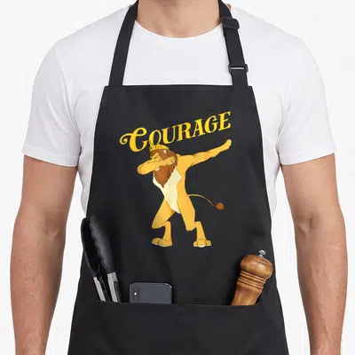 Dabbing lion apron - Cartoon lion in yellow and brown dabbing with arched COURAGE text above — shown on a black cotton canvas apron against a white background