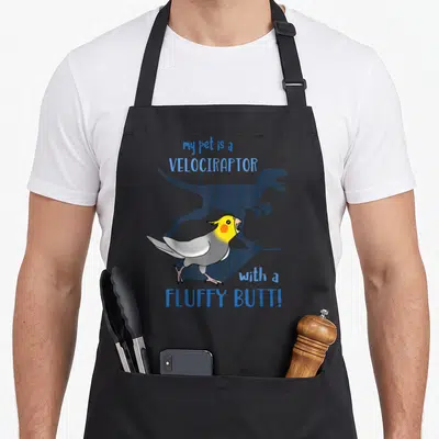 Velociraptor pet apron - Cartoon cockatiel standing before dark raptor shadow with blue text my pet is a VELOCIRAPTOR — shown on a black cotton canvas apron against a white background