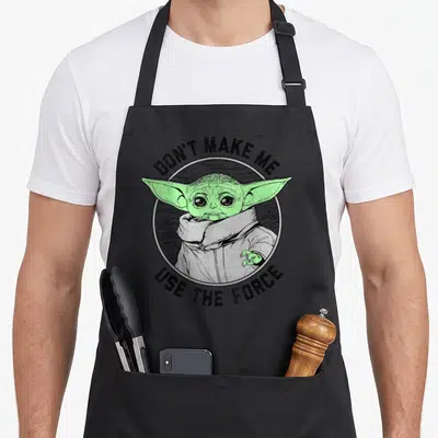 Funny force use of apron - Grogu in distressed circle with arched black text reading Don't Make Me Use The Force — shown on a black cotton canvas apron against a white background