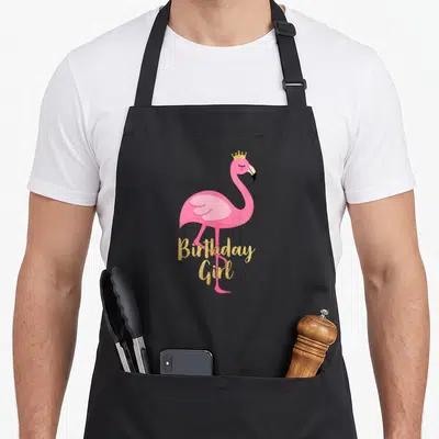 Birthday flamingo apron - Pink cartoon flamingo wearing a gold crown with gold glitter Birthday Girl text — shown on a black cotton canvas apron against a white background