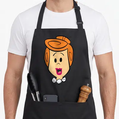 Wilma face apron - Cartoon woman's head with orange hair and pink lips on black fabric — shown on a black cotton canvas apron against a white background