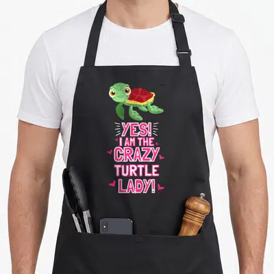 Outfit sea apron - Cartoon green turtle with red shell above pink text Yes I Am The Crazy Turtle Lady surrounded — shown on a black cotton canvas apron against a white background