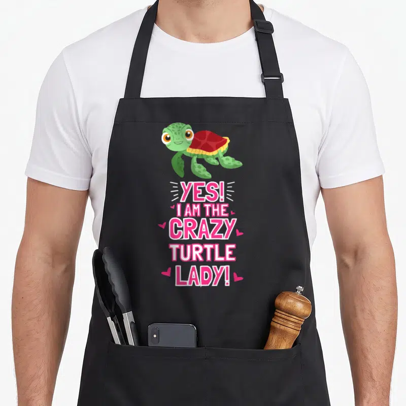 Outfit sea apron - Cartoon green turtle with red shell above pink text Yes I Am The Crazy Turtle Lady surrounded — shown on a black cotton canvas apron against a white background