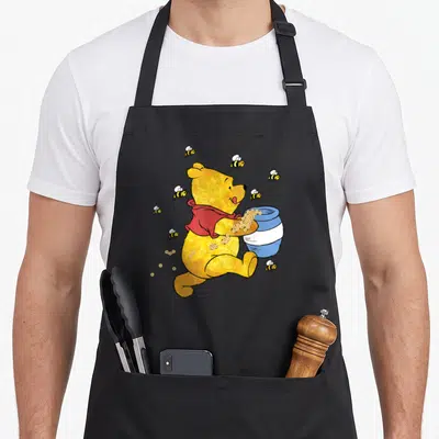 Jar honey bee apron - Yellow cartoon bear holding blue pot surrounded by bees in red and gold — shown on a black cotton canvas apron against a white background