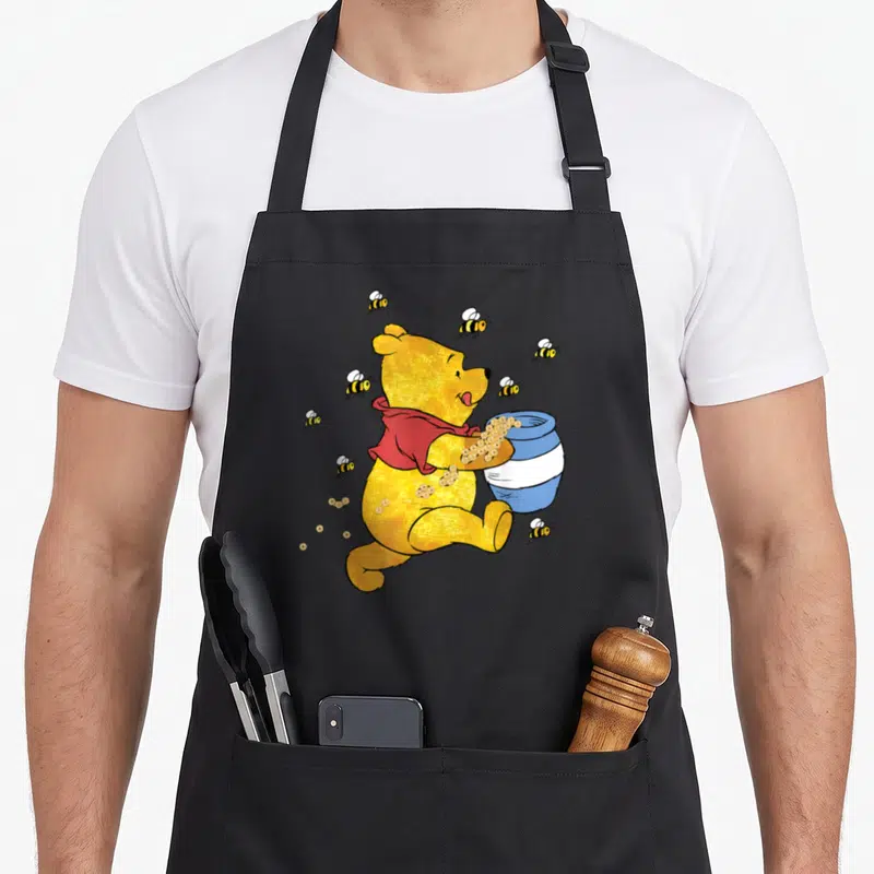 Jar honey bee apron - Yellow cartoon bear holding blue pot surrounded by bees in red and gold — shown on a black cotton canvas apron against a white background
