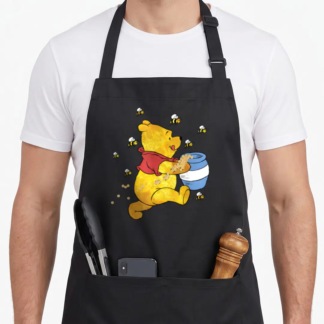 Jar honey bee apron - Yellow cartoon bear holding blue pot surrounded by bees in red and gold — shown on a black cotton canvas apron against a white background