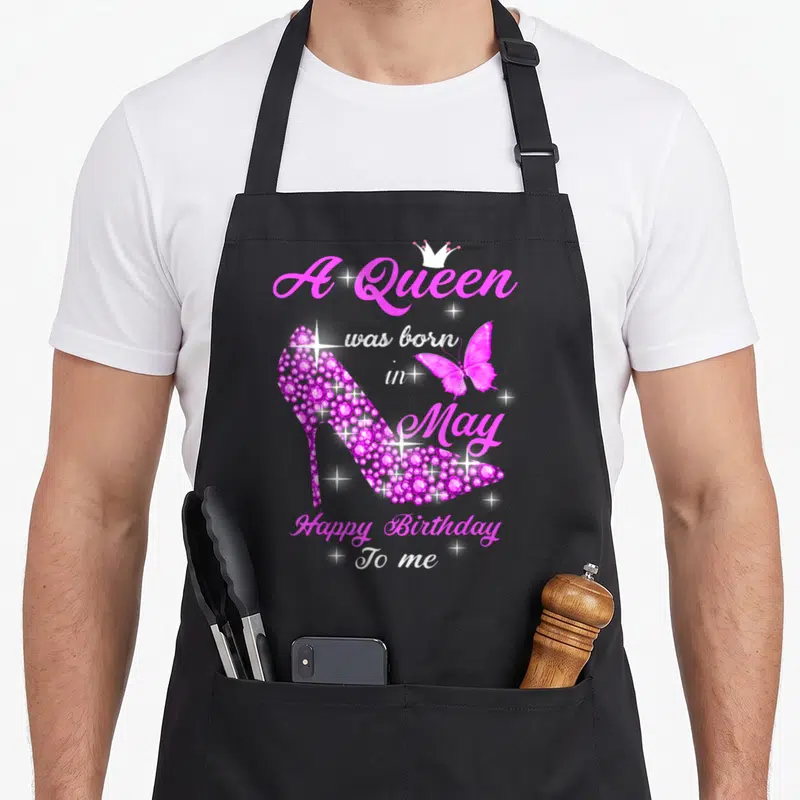 May baking queen apron - Pink rhinestone high heel shoe with butterfly and A Queen was born in May text — shown on a black cotton canvas apron against a white background
