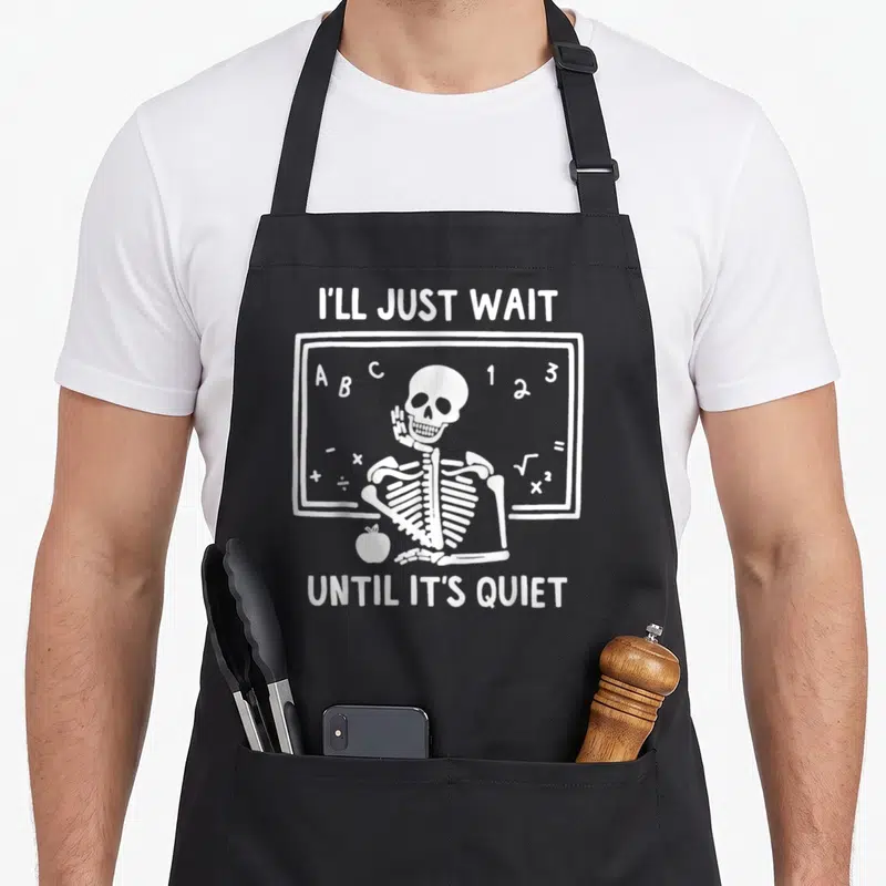 Ill skeleton apron - White cartoon skeleton sitting at a chalkboard with an apple and text reading I'll Just Wait Until — shown on a black cotton canvas apron against a white background