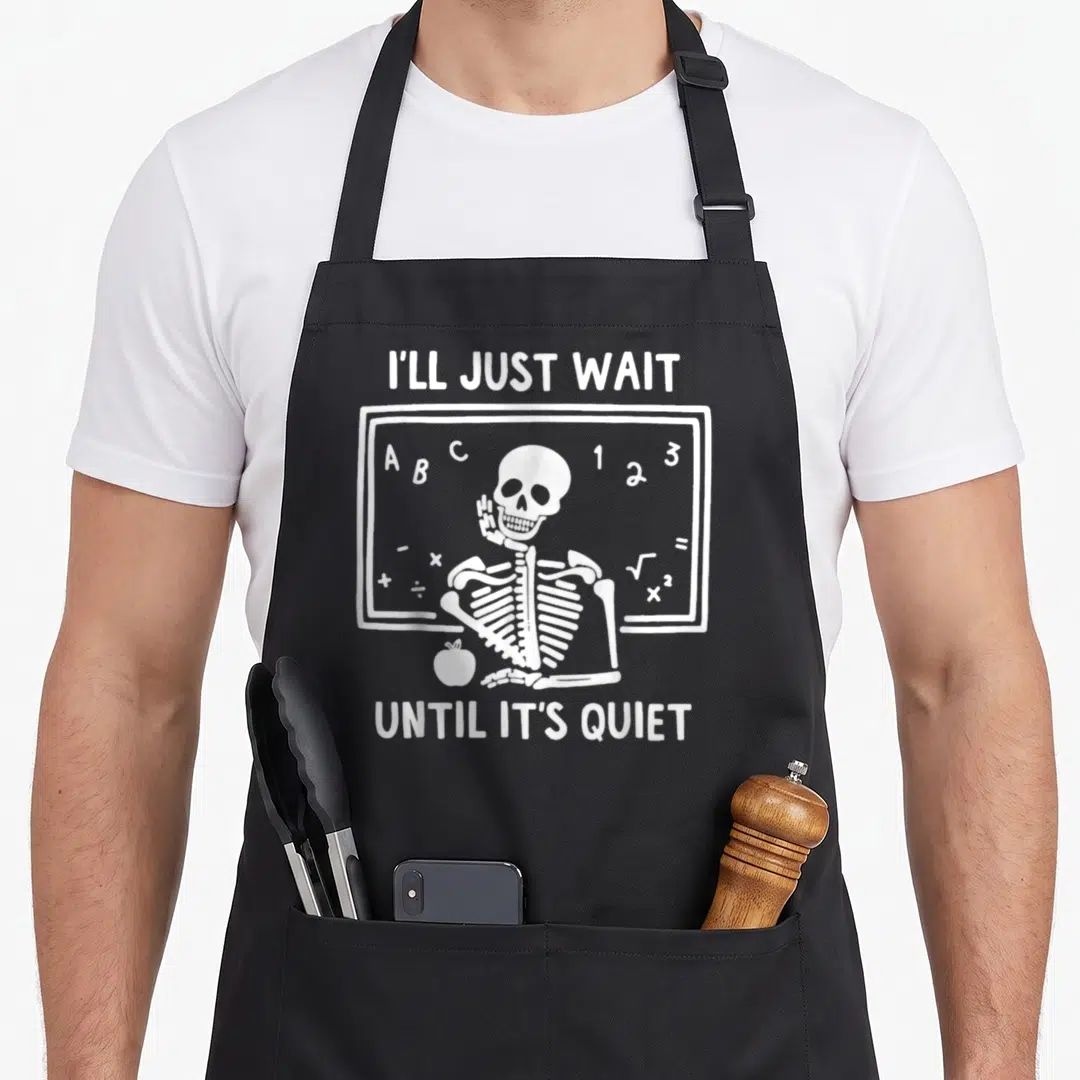 Ill skeleton apron - White cartoon skeleton sitting at a chalkboard with an apple and text reading I'll Just Wait Until — shown on a black cotton canvas apron against a white background