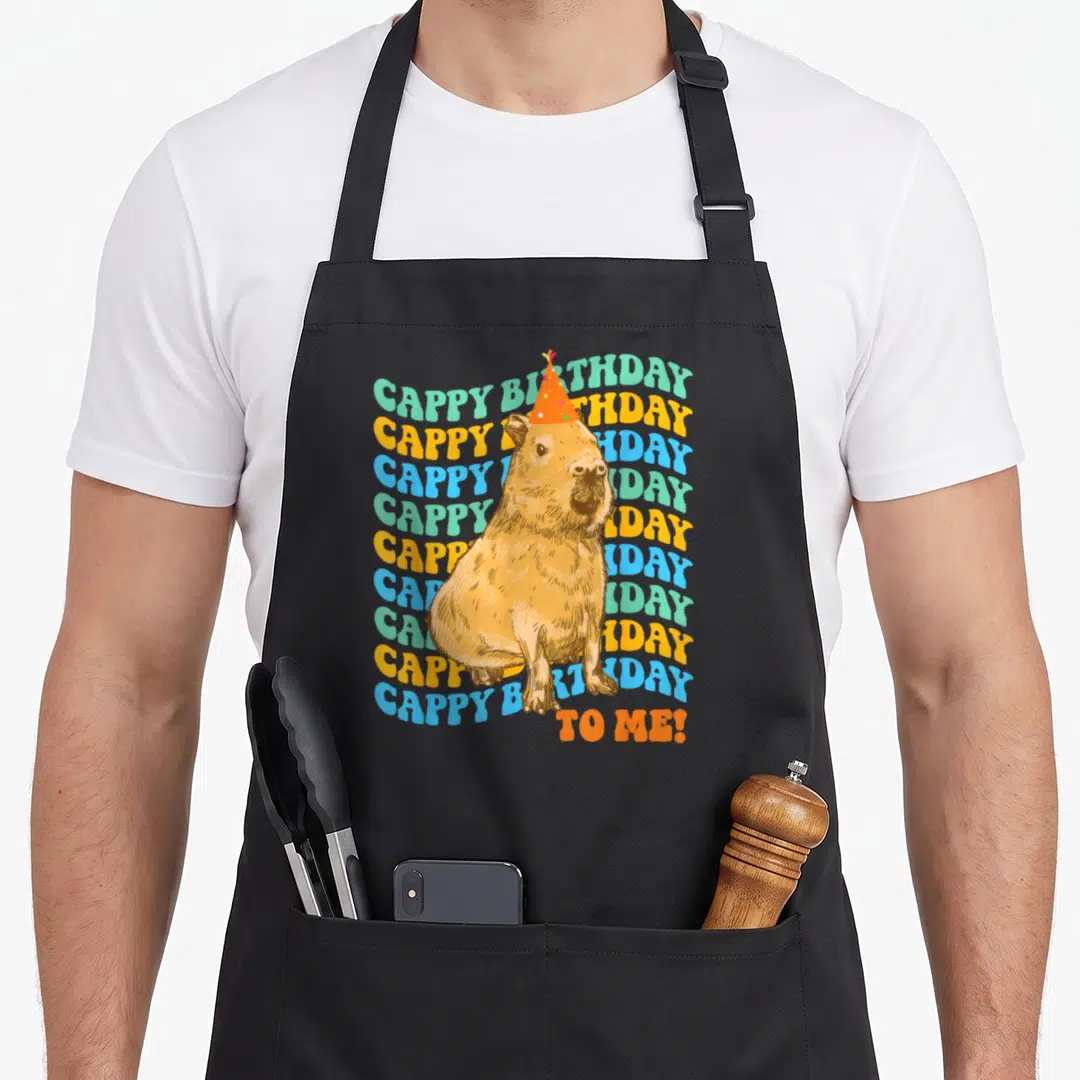 Cute capybara birthday apron - Brown capybara in orange party hat surrounded by ten wavy lines of mint green blue — shown on a black cotton canvas apron against a white background