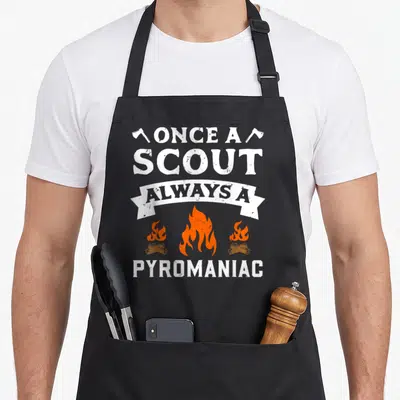 Once scouting apron - Distressed text once a scout always a pyromaniac with two axes, white ribbon banner, and three — shown on a black cotton canvas apron against a white background