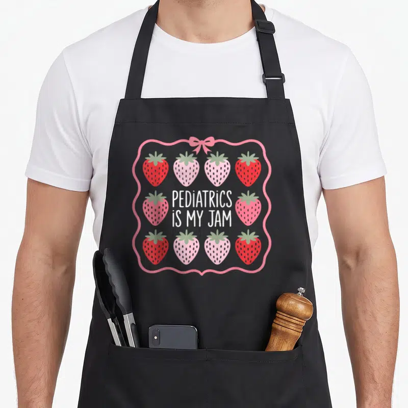 Pediatric strawberry apron - PEDIATRICS IS MY JAM text between two rows of six red and pink strawberries with pink bow — shown on a black cotton canvas apron against a white background