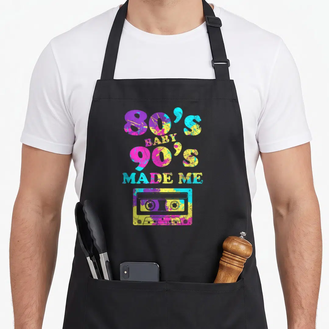 90s cassette 80s apron - Stacked '80'S BABY 90'S MADE ME' text above paint-splattered cassette in purple yellow blue — shown on a black cotton canvas apron against a white background