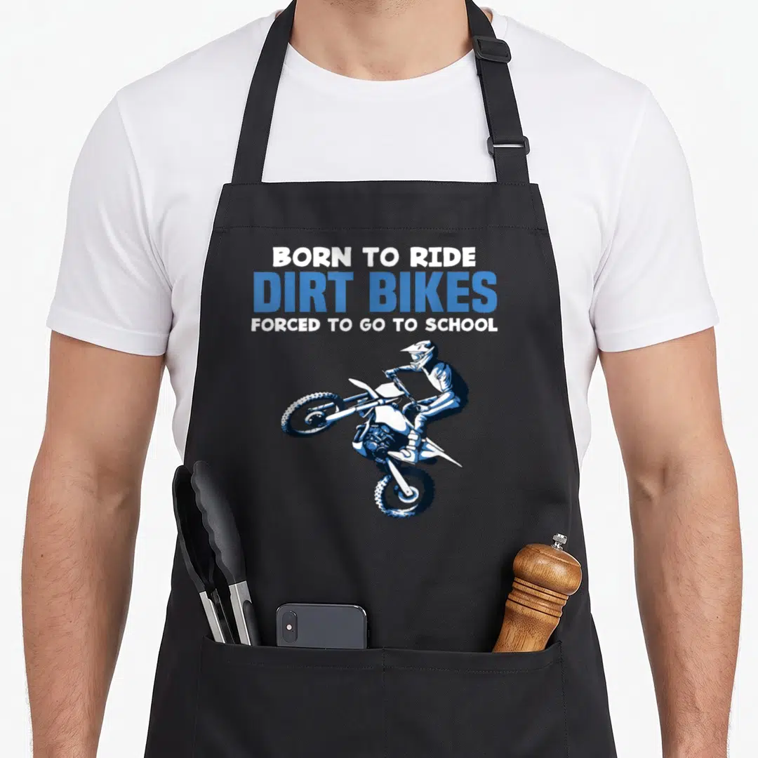Forced bike apron - BORN TO RIDE DIRT BIKES FORCED TO GO TO SCHOOL text above jumping cartoon rider — shown on a black cotton canvas apron against a white background