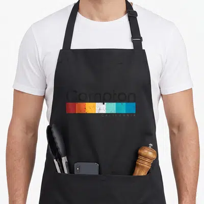 Vintage california compton apron - Compton text above seven distressed color blocks in red, orange, yellow, white — shown on a black cotton canvas apron against a white background