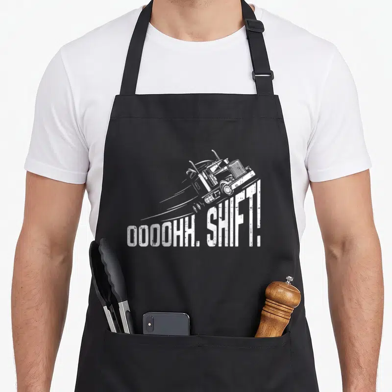Ohh semi truck apron - White line-art truck tilted above OOOOHH SHIFT text with speed lines on black fabric — shown on a black cotton canvas apron against a white background