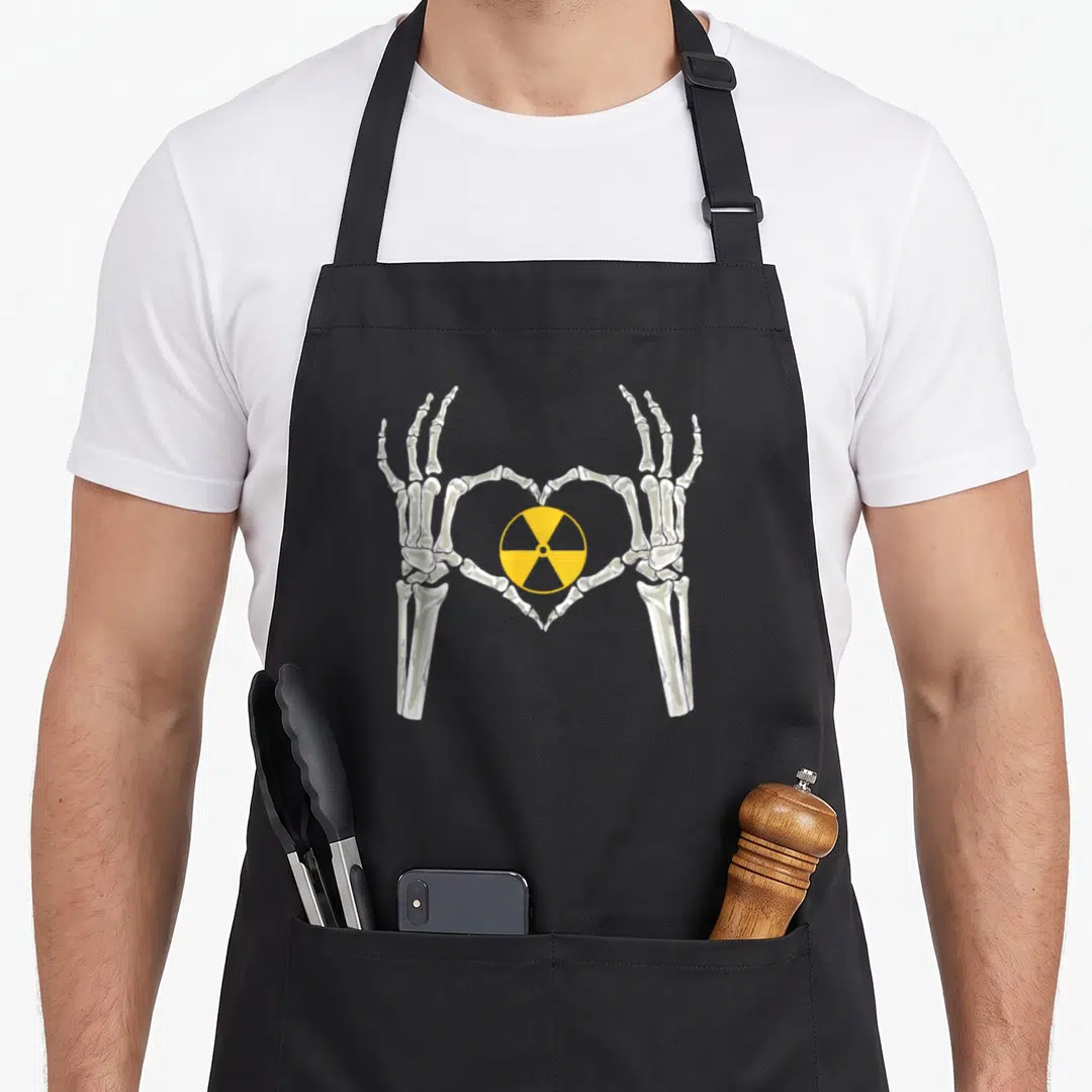 Rad skeleton apron - Two white skeleton hands form a heart enclosing a yellow radiation symbol on black fabric — shown on a black cotton canvas apron against a white background