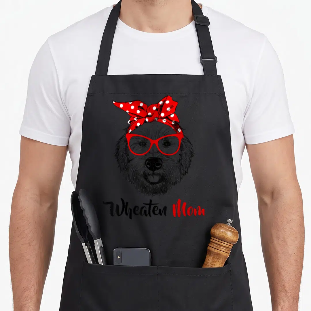 Wheaten dog apron - Cartoon dog head with red glasses and red polka dot bow, text Wheaten Mom below — shown on a black cotton canvas apron against a white background