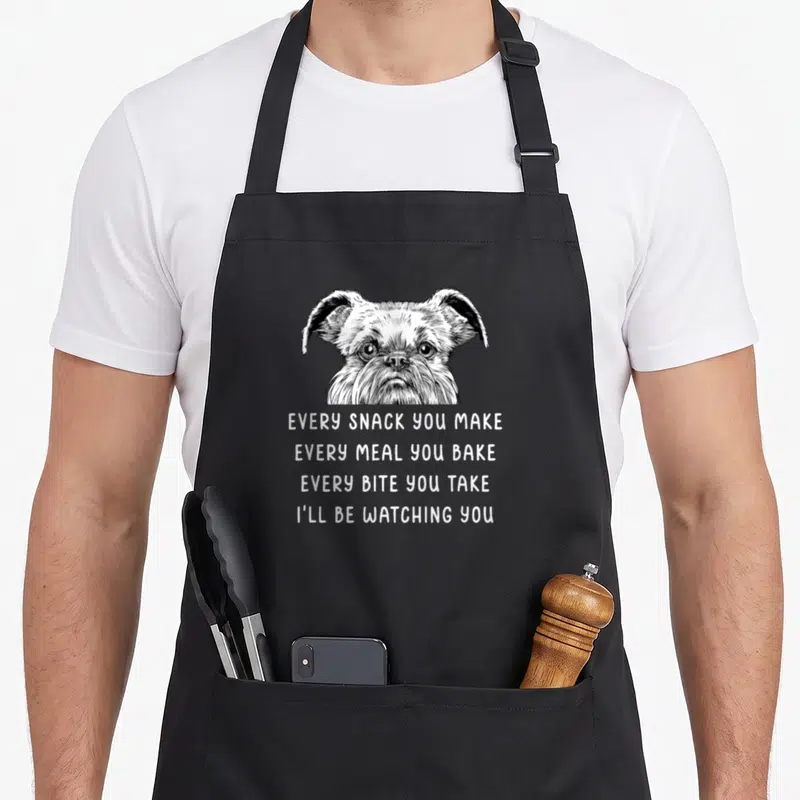 Snack brussels apron - White line art dog head above four lines of text reading I'll be watching you — shown on a black cotton canvas apron against a white background