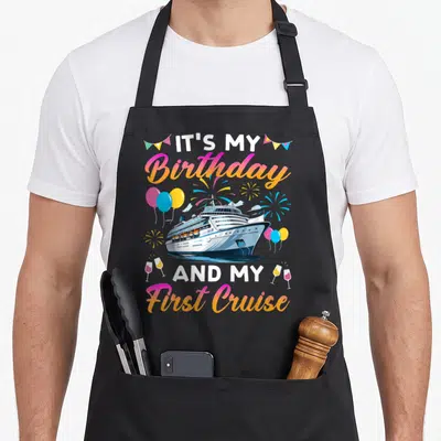 Cruise celebration birthday apron - Cartoon cruise ship with balloons, fireworks, wine glasses, and gradient text — shown on a black cotton canvas apron against a white background