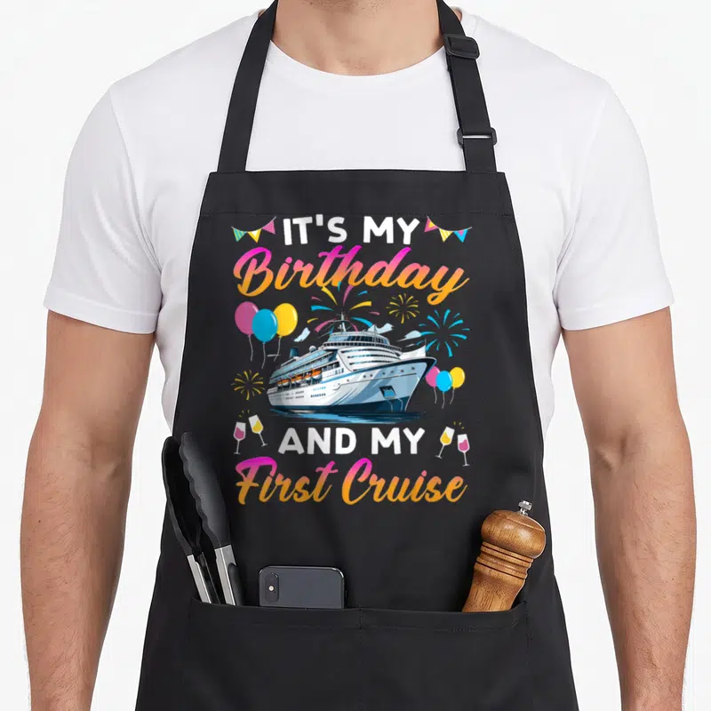 Cruise celebration birthday apron - Cartoon cruise ship with balloons, fireworks, wine glasses, and gradient text — shown on a black cotton canvas apron against a white background
