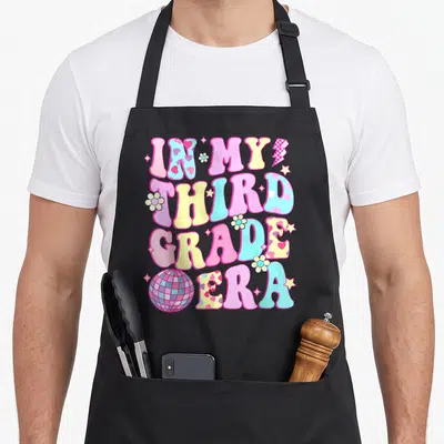 Groovy grade era apron - Bubbly text reading IN MY THIRD GRADE ERA with disco ball, flowers, and stars in pink yellow — shown on a black cotton canvas apron against a white background