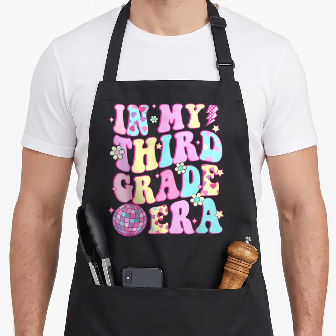 Groovy grade era apron - Bubbly text reading IN MY THIRD GRADE ERA with disco ball, flowers, and stars in pink yellow — shown on a black cotton canvas apron against a white background