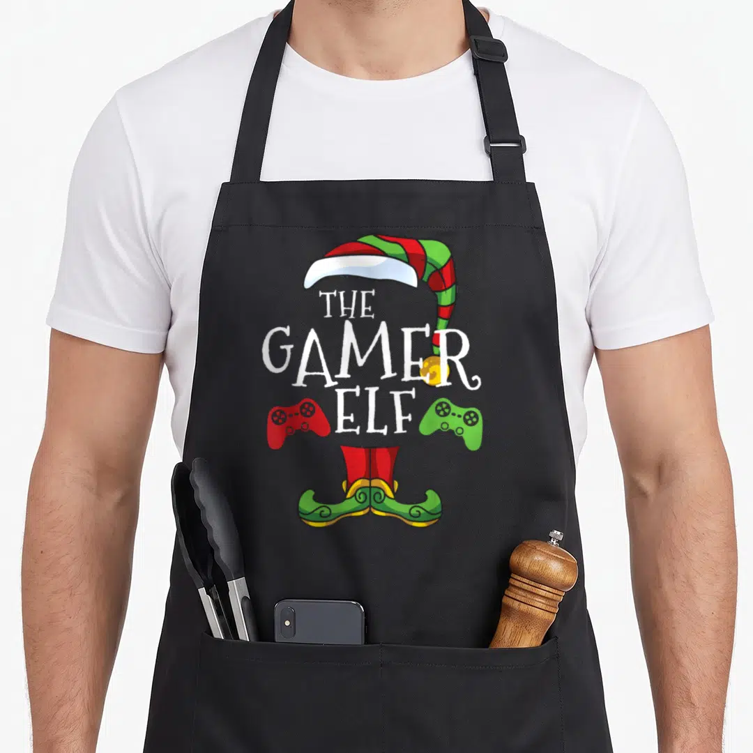 Video christmas elf apron - THE GAMER ELF text between red and green controllers with elf hat and shoes — shown on a black cotton canvas apron against a white background