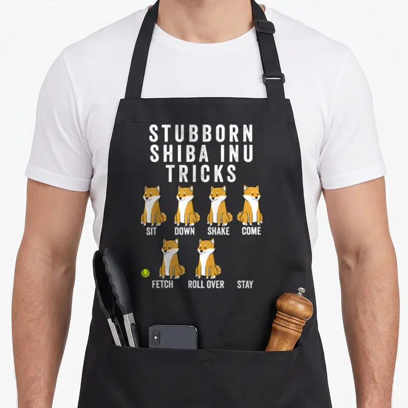 Stubborn dog apron - Six cartoon Shiba Inus in orange and white with trick names and a yellow-green tennis ball — shown on a black cotton canvas apron against a white background