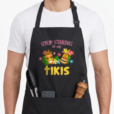 Stop tropical print aprons - Two cartoon tiki masks with pink flowers and leaves under text STOP STARING at my TIKIS — shown on a black cotton canvas apron against a white background