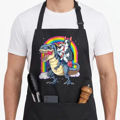 Terrier dinosaur apron - Boston Terrier riding white unicorn atop blue T-rex under rainbow arch with stars and clouds — shown on a black cotton canvas apron against a white background