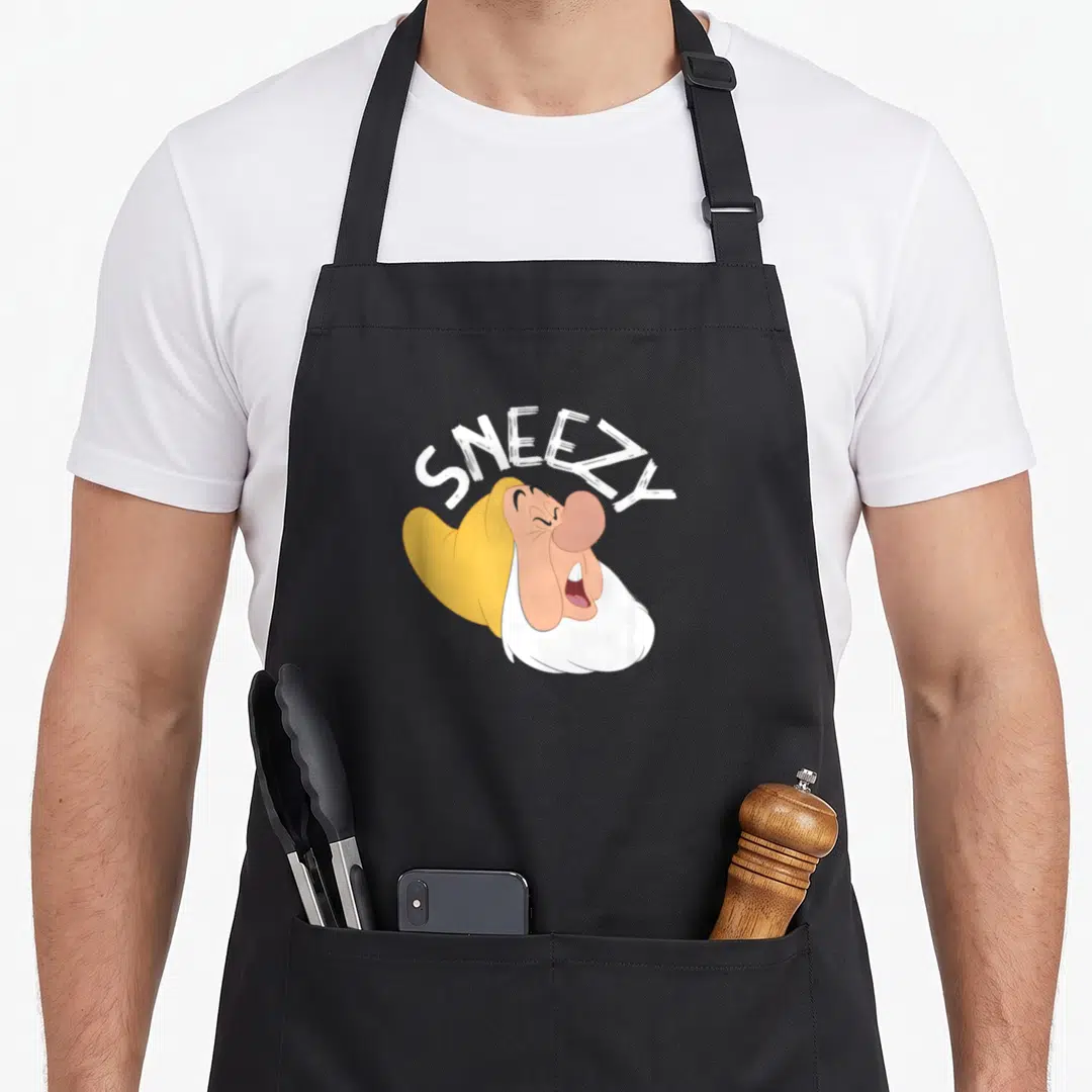 Sneezy snow white apron - Cartoon dwarf sneezing with SNEEZY text in yellow and white — shown on a black cotton canvas apron against a white background