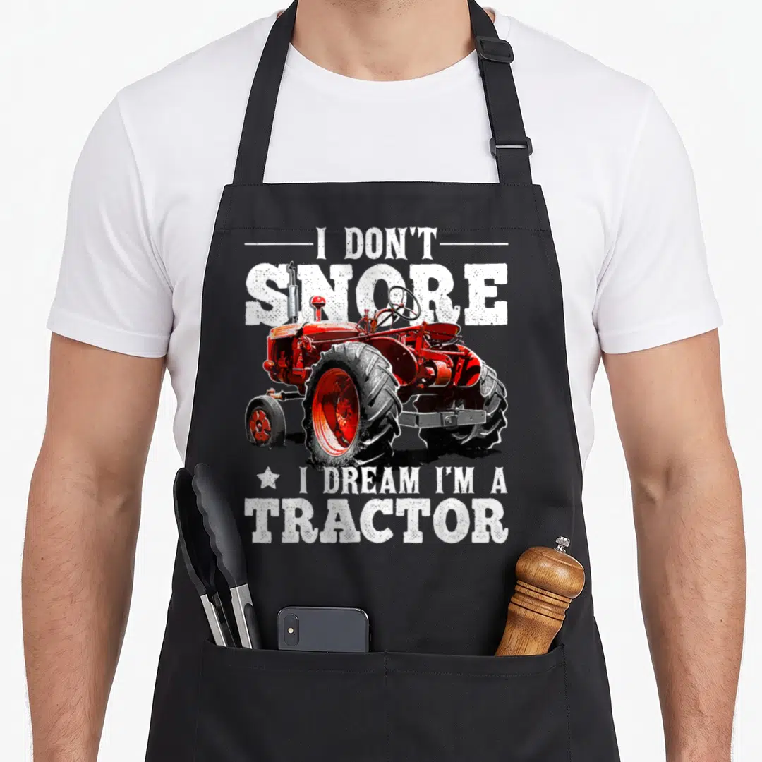 Snore farmer apron - Red tractor graphic with white distressed text I Don't Snore I Dream I'm a Tractor — shown on a black cotton canvas apron against a white background