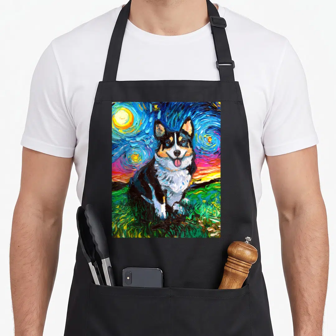 Starry corgi aprons - Tri-color corgi sitting on green grass under a swirling blue and yellow starry night sky — shown on a black cotton canvas apron against a white background
