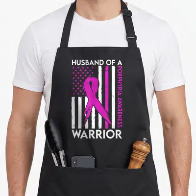 Porphyria husband apron - Pink awareness ribbon over distressed USA flag with HUSBAND OF A WARRIOR text — shown on a black cotton canvas apron against a white background