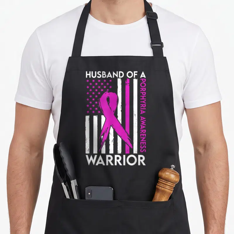 Porphyria husband apron - Pink awareness ribbon over distressed USA flag with HUSBAND OF A WARRIOR text — shown on a black cotton canvas apron against a white background