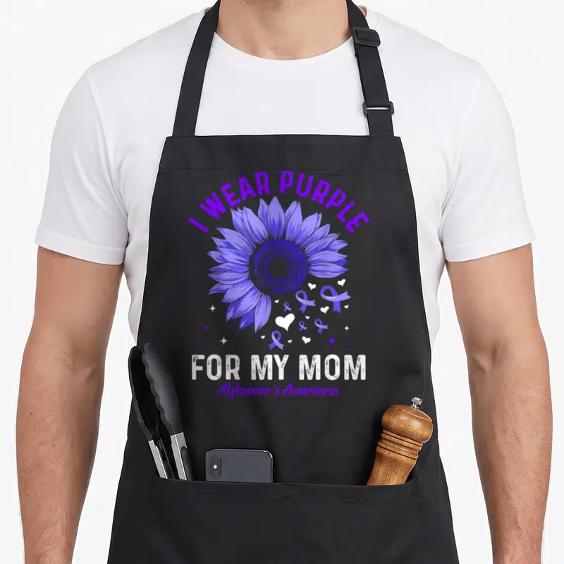 Vintage dementia alzheimer's aprons - Purple sunflower with I WEAR PURPLE text and scattered ribbons hearts stars — shown on a black cotton canvas apron against a white background