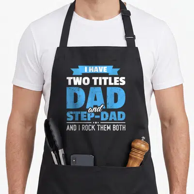 Title step father day apron - Blue and white typography reading I have two titles dad and step-dad with ribbon and stars — shown on a black cotton canvas apron against a white background