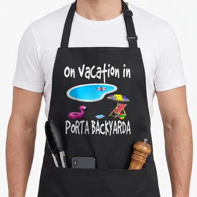 Staycation flamingo apron - Cartoon blue pool with pink flamingo float and text on Vacation in PORTA BACKYARDA — shown on a black cotton canvas apron against a white background