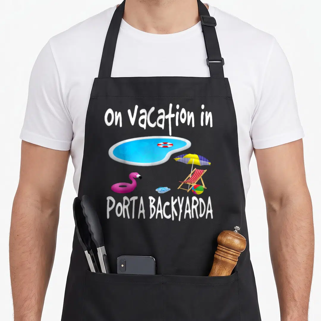 Staycation flamingo apron - Cartoon blue pool with pink flamingo float and text on Vacation in PORTA BACKYARDA — shown on a black cotton canvas apron against a white background