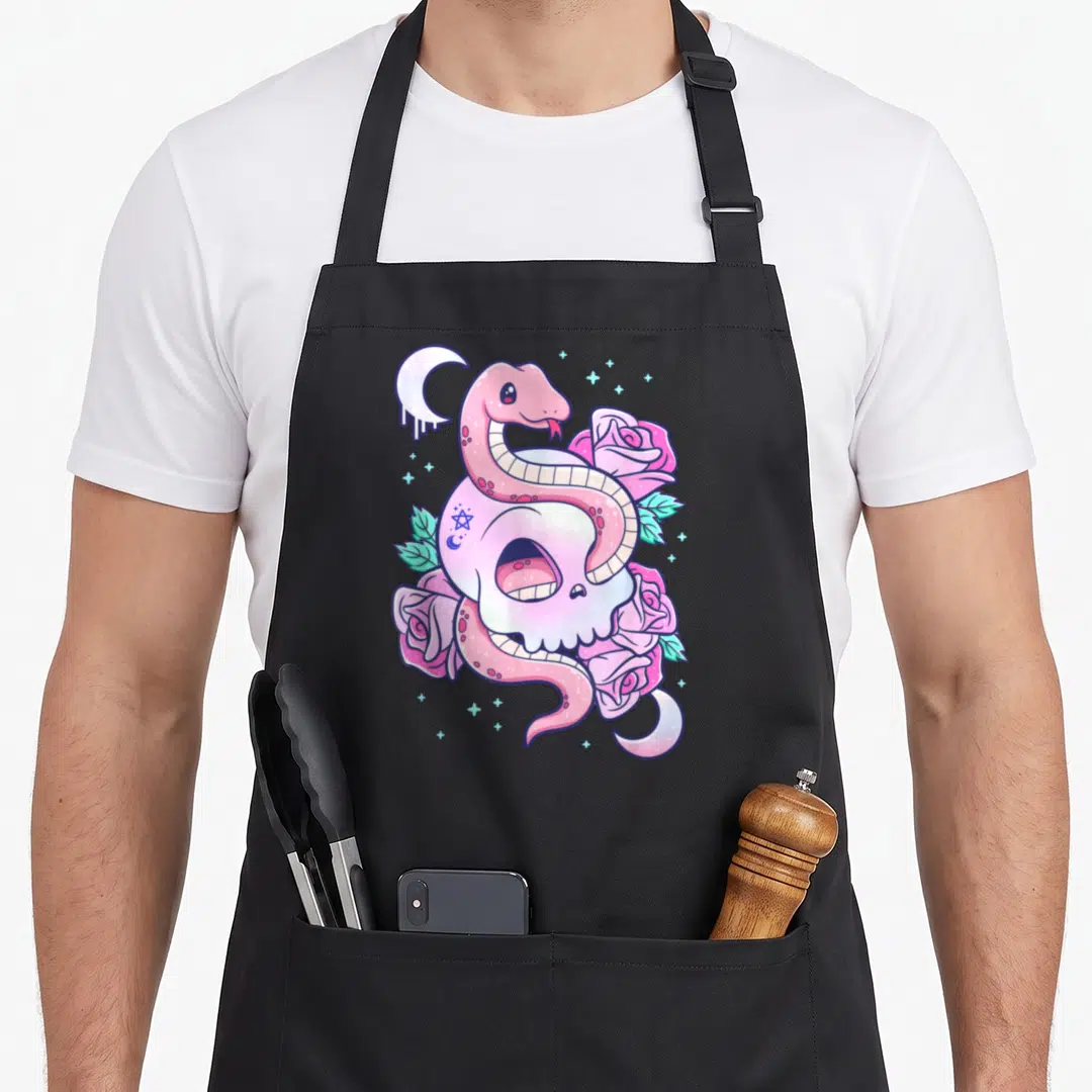 Scary goth apron - Pink snake coiled around white skull with roses and moons in cartoon style — shown on a black cotton canvas apron against a white background